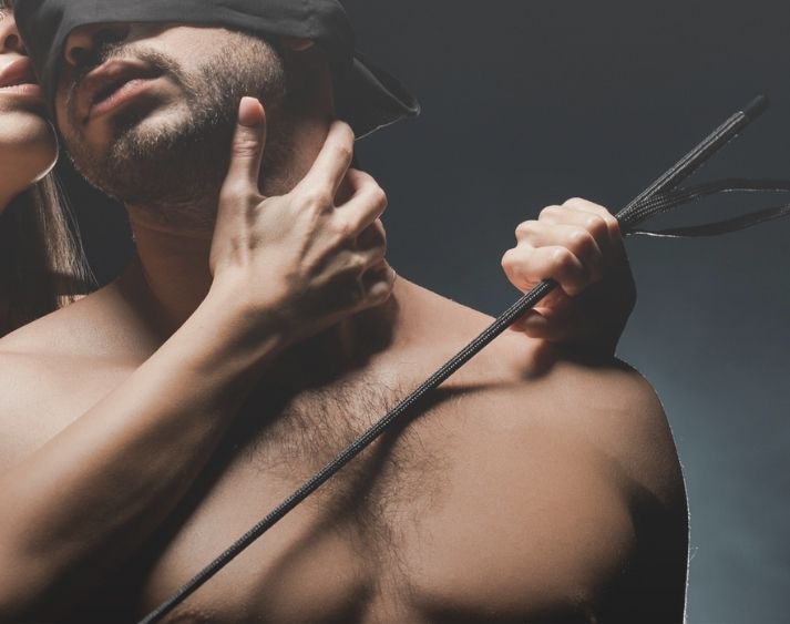Dominant female embracing blindfolded male with a riding crop adult toy